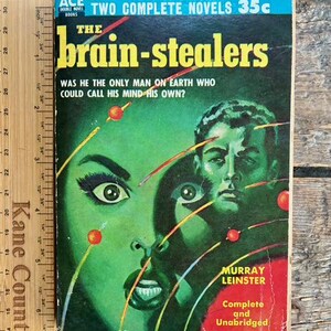 May include: Vintage paperback book titled "The Brain-Stealers" with a green and red cover. The cover features a woman's face and a man's face, with yellow text and the author's name, Murray Leinster. The book is from Ace Double Books.