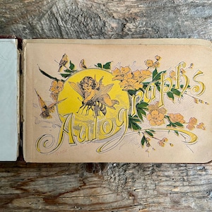 Pode incluir: Livro de autógrafos antigo aberto com uma capa decorativa. A capa apresenta a palavra "Autographs" em letras ornamentadas, com uma ilustração de querubim e abelha em um círculo amarelo, rodeada por flores e pássaros.