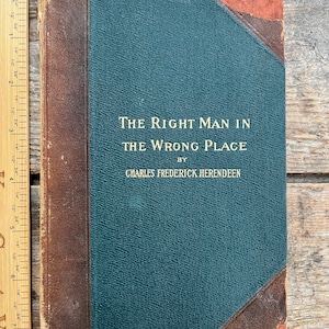 May include: An antique book titled "The Right Man in the Wrong Place" by Charles Frederick Herendeen. The book has a teal cover with brown leather corners and spine. A wooden ruler is placed next to the book.