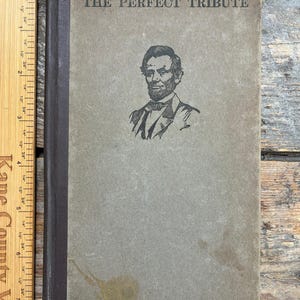 May include: Vintage book titled "The Perfect Tribute" with a portrait of Abraham Lincoln. The cover is a faded gray with dark brown spine. The author's name, Mary Raymond Shipman Andrews, is printed at the bottom.