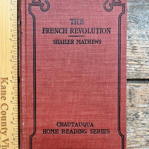 May include: A vintage book titled "The French Revolution" by Shailer Mathews. The book has a red cover with black decorative borders and text. The book is part of the Chautauqua Home Reading Series. A wooden ruler is on the left side.