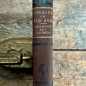 May include: A brown antique book spine with black decorative accents and gold lettering that reads Diseases of Old Age, Charcot and Loomis. The spine features a palm-like botanical motif and horizontal lines, showing signs of age and wear.