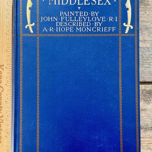 May include: A vintage book titled "Middlesex" with a blue cover and gold lettering. The cover features decorative gold accents and the names of the author and illustrator. The book is displayed on a wooden surface.