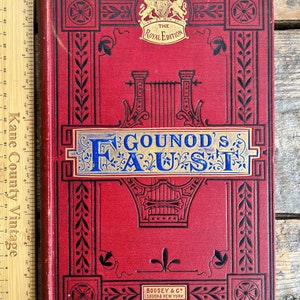 Może przedstawiać: Czerwona książka w twardej oprawie z pozłacanymi napisami zatytułowana "Gounod's Faust" z herbem królewskim i napisem "The Royal Edition" na okładce. Książka jest ozdobiona czarnymi i złotymi zdobieniami.