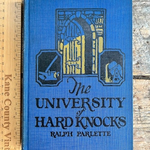 Könnte beinhalten: Vintage Hardcover-Buch mit dem Titel "The University of Hard Knocks" von Ralph Parlette. Der blaue Einband zeigt eine schwarz-goldene Illustration eines gewölbten Eingangs und anderer architektonischer Elemente. Das Buch ist etwa 23 cm hoch.