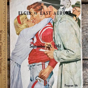 Könnte beinhalten: Ein Vintage-Programmcover für ein Footballspiel zwischen Elgin und East Aurora am 29. September 1961. Das Cover zeigt ein Paar, das sich küsst, wobei der Mann ein rot-weißes Footballtrikot trägt und die Frau ein weißes Kleid. Das Programm kostet 10 Cent.
