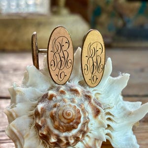 May include: A pair of oval-shaped gold-toned cufflinks featuring an intricate, dark-colored engraved monogram of the letters ARB. The cufflinks have a polished metallic finish and are displayed upright, showcasing the detailed script lettering.
