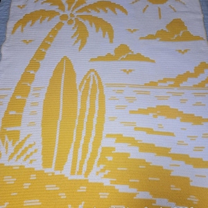 Peut inclure: Une couverture crochetée blanche et jaune représentant une scène de plage avec un palmier, deux planches de surf, un soleil, des nuages et des oiseaux. Le texte "Crocheted by Marie Sparks · 2026" est intégré dans le motif en bas à droite.