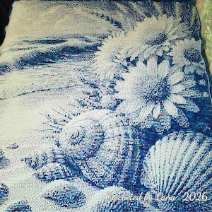 May include: A rectangular crochet blanket featuring a blue and white floral pattern with large daisy-like flowers. The textile displays a gradient of blue shades and includes the text Crocheted by Lana 2026 in the bottom right corner.