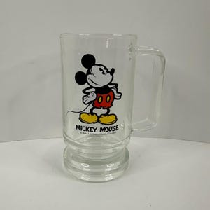 May include: A clear glass mug featuring a graphic of Mickey Mouse with black ears, red shorts, and yellow shoes. The text MICKEY MOUSE is printed in black below the character, with the smaller text WALT DISNEY PRODUCTIONS underneath it.
