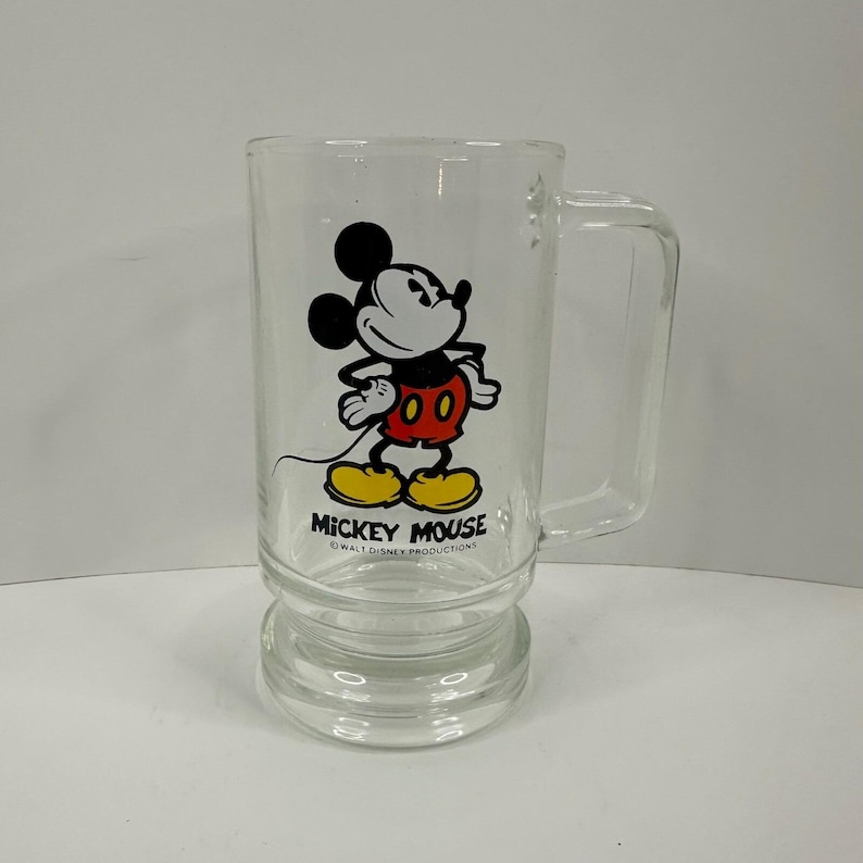 May include: A clear glass mug featuring a graphic of Mickey Mouse with black ears, red shorts, and yellow shoes. The text MICKEY MOUSE is printed in black below the character, with the smaller text WALT DISNEY PRODUCTIONS underneath it.