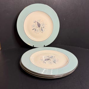 May include: A set of round dinner plates featuring a wide, pale mint green border and a cream-coloured centre. The centre of each plate is decorated with a delicate floral illustration in shades of grey and blue, accented by small red dots.