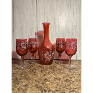 May include: A vintage red glass wine decanter and four matching wine glasses. The set features a grape design in silver. The glasses and decanter are on a brown surface with a light wood background.