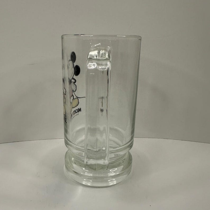 May include: A clear glass mug featuring a graphic of Mickey Mouse on the side. The mug has a sturdy handle and a thick, reinforced base. The text MICKEY MOUSE is printed in black near the character graphic on the side of the glass vessel.