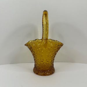May include: Amber-colored glass basket with a handle. The basket has a textured diamond pattern and a scalloped rim. The handle is arched and attached to the sides of the basket. A decorative piece.