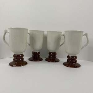 May include: Four white ceramic mugs with brown pedestal bases and handles. The mugs have a classic design, suitable for coffee or tea. The bases have a stacked, rounded design.