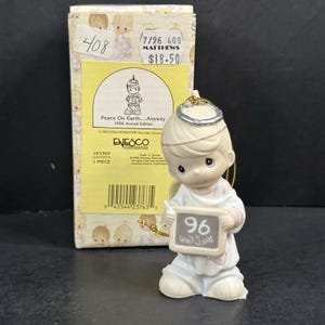 May include: A Precious Moments porcelain ornament featuring a child with a silver halo holding a sign that reads 96. The packaging box is light yellow and white with the text Peace On Earth...Anyway 1996 Annual Edition and Enesco Corporation.
