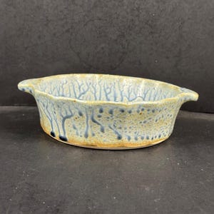 May include: A shallow, oval ceramic bowl with two small handles on opposite sides. The exterior and interior feature a textured, speckled beige glaze with blue, tree-like drip patterns running vertically down the sides of the vessel.