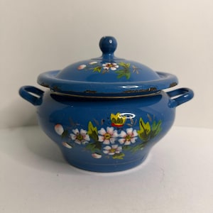 Könnte beinhalten: Eine blaue Emaille-Schüssel mit Deckel und zwei seitlichen Griffen, verziert mit handgemalten weißen Blumen mit gelben Mitten, grünen Blättern und einem kleinen gelben Schmetterling. Der Rand des Deckels zeigt Abnutzungserscheinungen mit dunklem Metall.