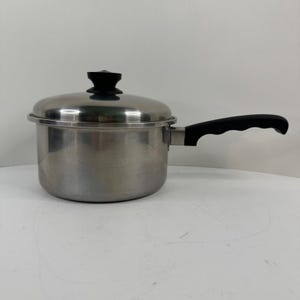 Op de afbeelding: Een roestvrijstalen steelpan met bijpassend deksel. De pan heeft een cilindrische vorm, een enkele zwarte hittebestendige handgreep met een voorgevormde grip en een zwarte knop in het midden van het deksel. Het metalen oppervlak is gepolijst.