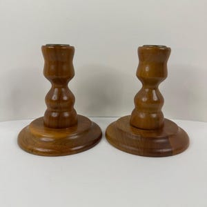 May include: Two wooden candle holders. Each holder has a round base and a turned, bulbous stem. The wood has a warm, medium brown tone. The top of each holder is a small, flat surface, likely to hold a candle. The holders are displayed on a white surface.