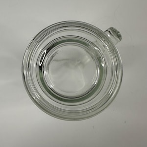 May include: A clear glass mug viewed from directly above, showcasing a circular rim, a smooth interior base, and a thick glass wall structure. A small portion of the handle is visible extending from the right side of the circular vessel.