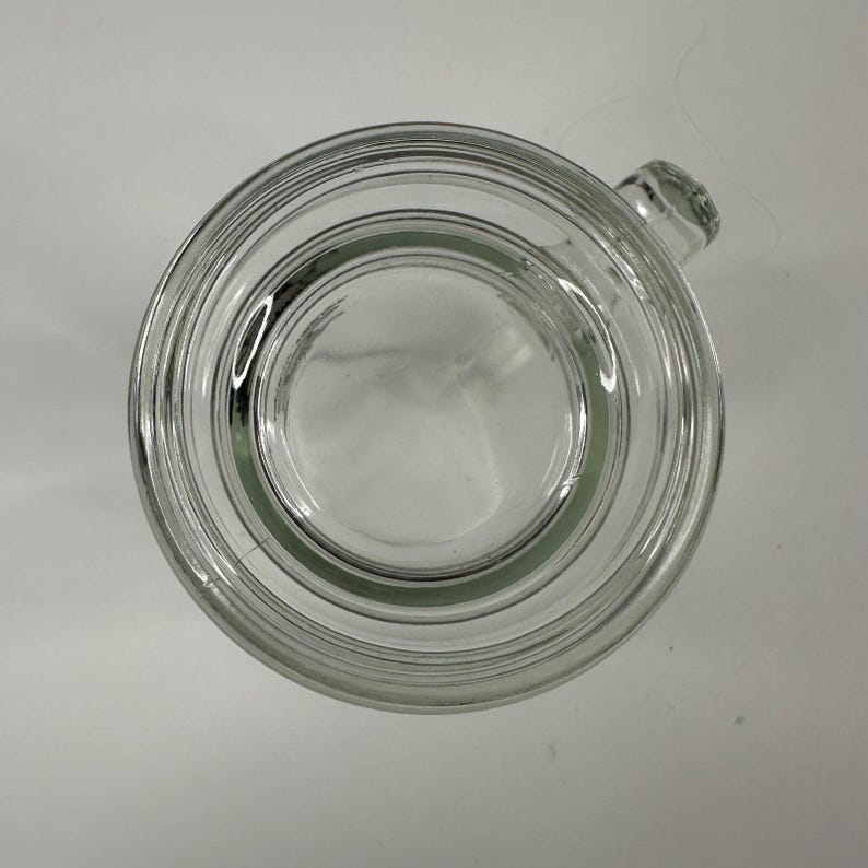 May include: A clear glass mug viewed from directly above, showcasing a circular rim, a smooth interior base, and a thick glass wall structure. A small portion of the handle is visible extending from the right side of the circular vessel.