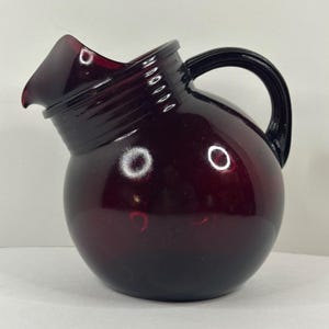 May include: A vintage, ruby-red glass pitcher with a rounded body and a curved spout. The pitcher features a black handle and a ribbed design around the neck. The glass has a glossy finish, reflecting light. A classic piece for serving beverages.