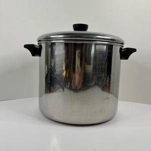 Op de afbeelding: Een cilindrische roestvrijstalen soeppan met een gepolijste afwerking, voorzien van twee zwarte kunststof handgrepen aan de zijkanten en een bijpassende zwarte knop op het deksel. Het metalen oppervlak is reflecterend en glanzend.