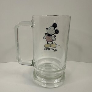 May include: A clear glass mug featuring a graphic of Mickey Mouse standing with hands on hips. The character wears pink shorts and yellow shoes. Below the illustration, the text reads MICKEY MOUSE and WALT DISNEY PRODUCTIONS in black.