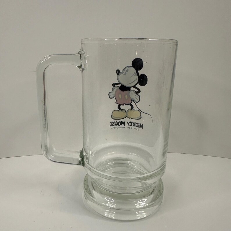 May include: A clear glass mug featuring a graphic of Mickey Mouse standing with hands on hips. The character wears pink shorts and yellow shoes. Below the illustration, the text reads MICKEY MOUSE and WALT DISNEY PRODUCTIONS in black.