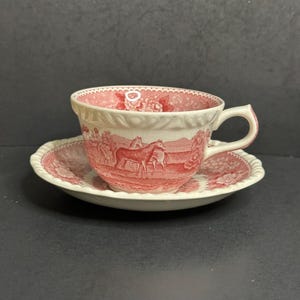 May include: A vintage teacup and saucer set. The white ceramic cup and saucer feature a red transferware design of horses in a landscape. The cup has a scalloped rim and handle. The saucer also has a scalloped edge.