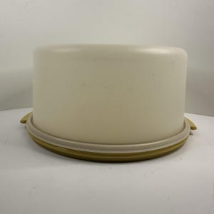 Puede incluir: Un portatartas de plástico blanco roto vintage con tapa a juego. La base es de color amarillo verdoso apagado, con un anillo beige claro y dos asas laterales. La tapa tiene forma de cilindro redondeado.