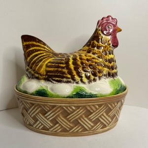May include: A ceramic hen-shaped lidded container featuring a brown and yellow feathered pattern, a red comb, and a yellow beak. The base is designed to resemble a woven basket, with the hen resting on a bed of white eggs and green grass.