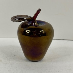 May include: A decorative glass apple sculpture with a bronze-colored body and a dark red stem and leaf. The apple has a glossy finish and a subtle iridescent sheen, adding depth and visual interest to the piece.