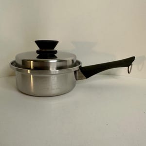 Puede incluir: Una cacerola de acero inoxidable con tapa y asa negra. La olla tiene marcas de medición en el lateral y un pomo negro en la tapa. El asa tiene un lazo en el extremo.