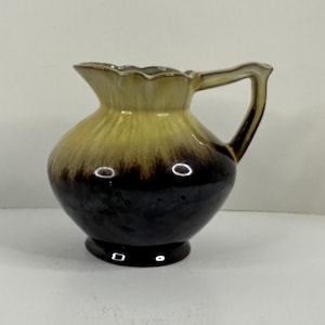 May include: A small, decorative ceramic jug with a handle. The jug has a rounded body that graduates from dark brown to yellow. The rim has a scalloped edge. The handle is a dark brown colour.