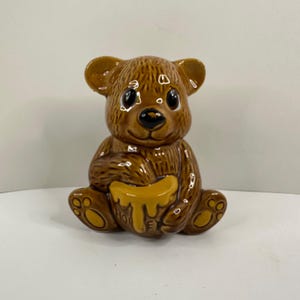 Puede incluir: Figura de oso de cerámica marrón sosteniendo un tarro de miel amarillo. El oso tiene ojos negros y una expresión sonriente. La palabra "HONEY" es visible en el tarro. La figura está sentada con las piernas cruzadas.