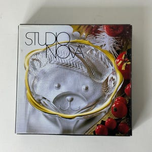 May include: A Studio Nova glass serving bowl in the shape of a teddy bear's face. The bowl has a gold rim and is presented in a box with the Studio Nova logo.