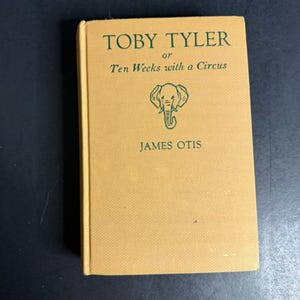 Pode incluir: Um livro de capa dura com uma capa amarela texturizada apresentando texto verde que diz Toby Tyler or Ten Weeks with a Circus de James Otis. Um desenho de linha verde de uma cabeça de elefante está centralizado entre o título e o autor.