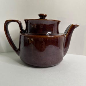 Può includere: Una teiera in ceramica marrone con coperchio e manico curvo. La teiera ha un beccuccio e un corpo arrotondato. La glassa ha una finitura lucida. La teiera è progettata per preparare il tè.