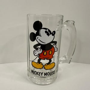 Peut inclure: Une chope &agrave; bi&egrave;re en verre transparent orn&eacute;e d'une illustration de Mickey Mouse avec des oreilles noires, un short rouge et des chaussures jaunes. Le texte MICKEY MOUSE est imprim&eacute; en lettres capitales noires sous le personnage, avec Disney.