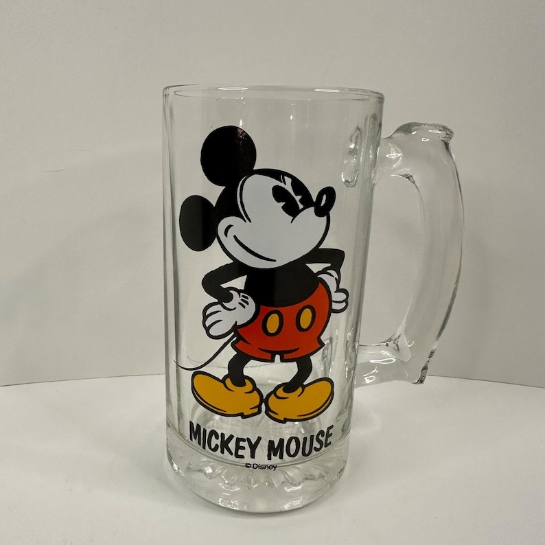 Peut inclure: Une chope &agrave; bi&egrave;re en verre transparent orn&eacute;e d'une illustration de Mickey Mouse avec des oreilles noires, un short rouge et des chaussures jaunes. Le texte MICKEY MOUSE est imprim&eacute; en lettres capitales noires sous le personnage, avec Disney.
