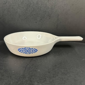 Puede incluir: Sartén de cerámica blanca con un mango largo. La sartén tiene un diseño decorativo azul y blanco en el lateral. La sartén está vacía y parece sin usar. La sartén está sobre una superficie oscura.