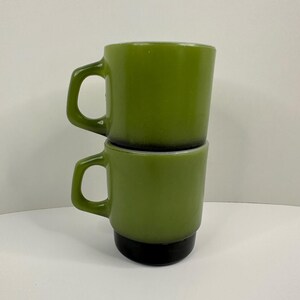 May include: Two stacked green mugs with black bases and curved handles. The mugs have a glossy finish and a simple, classic design. The mugs are empty and ready to be filled with your favorite beverage.