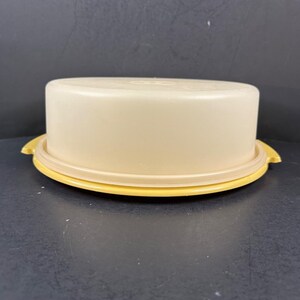 Puede incluir: Una tapa redonda de plástico beige para recipiente de alimentos con una parte superior lisa y plana y un borde ligeramente elevado. La tapa presenta una base circular de color amarillo brillante con dos pequeñas asas que sobresalen.
