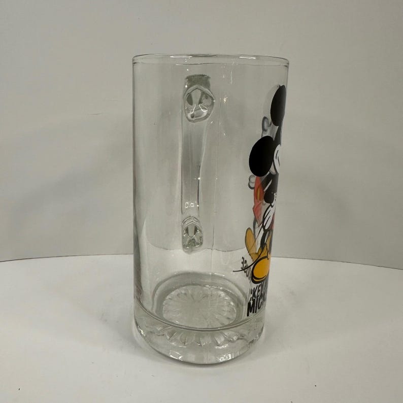 Peut inclure: Une chope &agrave; bi&egrave;re en verre transparent orn&eacute;e d'un motif de Mickey Mouse aux couleurs noir, blanc, rouge et jaune. La chope poss&egrave;de une anse robuste sur le c&ocirc;t&eacute; et une base textur&eacute;e. Le texte Mickey Mouse est imprim&eacute; verticalement en noir.