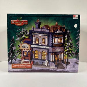 May include: A Coventry Cove porcelain lighted house in a box. The building features a blue and grey facade with gold accents, labelled Police and Village Tailor. It includes a 183 cm cord, illuminated windows, and a sign dated 1902 on the exterior.