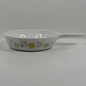 May include: White ceramic skillet with a long handle. The skillet is decorated with a floral pattern in yellow, blue, and white. The skillet is designed for cooking and serving food.