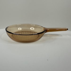 May include: A round, amber-coloured glass skillet with a smooth, transparent finish and a single integrated handle extending from the side. The base of the interior features a subtle, circular grid pattern embossed into the glass surface.